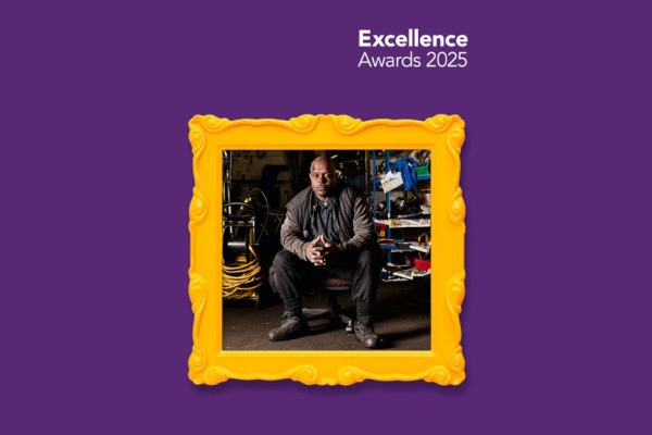 Superimposed on a purple background is a gold picture frame. Inside the frame is a photograph of a black man in black clothing sat in a workshop surrounded by tools and equipment.